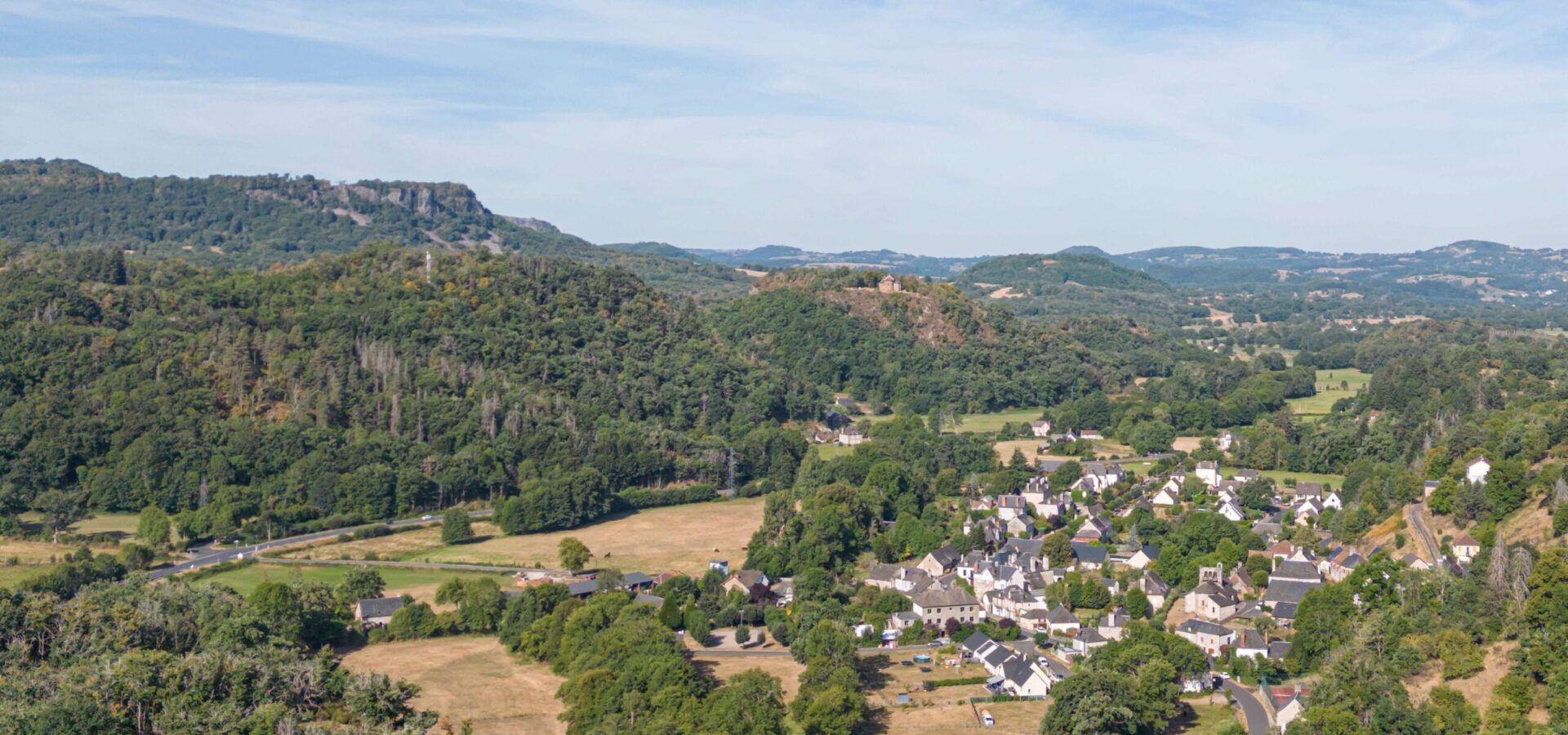 Vue de haut du village d'Antignac avec Plateau de Chastel Malhac au loin, sous un grand ciel bleu et végétation verdoyante.